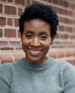 Smiling Black Woman with Short Curly Hair Wearing a Gray Knit Sweater, Leaning Against a Brick Wall in the Background.