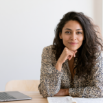 Woman with Curly Hair Sits at a Desk with an Open Laptop and Notebook, Resting Chin on Her Hand and Smiling in a Bright Room.