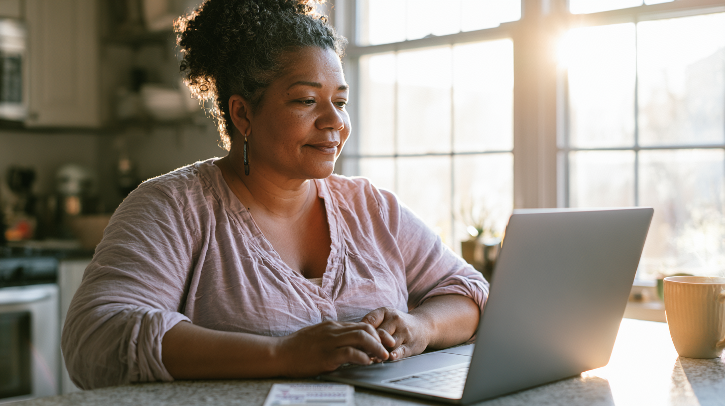Woman in a Pink Blouse Typing on a Laptop at a Sunlit Kitchen Counter, Mug Nearby.