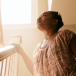 anxiety-ruining-my-life - Caring Clarity Counseling Woman on a Staircase, Gripping the Wooden Handrail and Looking Up Toward a Bright Window.