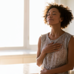 anxiety-soothing-techniques - Caring Clarity Counseling Woman with Curly Hair in a Sleeveless Top Stands in a Sunlit Kitchen, Eyes Closed, Hand on Her Chest in a Moment of Calm.