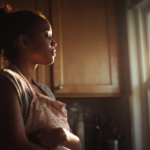 bipolar-and-postpartum-depression-1 - Caring Clarity Counseling Woman in a Kitchen Wearing an Apron Stands in Profile As Warm Sunlight Pours in from a Window, Eyes Closed in a Calm Moment.