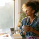 cbt-for-postpartum-depression - Caring Clarity Counseling Woman with Curly Hair in a Denim Shirt Sits by a Sunlit Kitchen Window, Holding a Mug and Looking Contemplatively Outside.