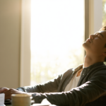 cigna-therapist-jersey-city-nj - Caring Clarity Counseling Man Seated at a Desk with a Laptop, Eyes Closed and Head Tilted Back As Sunlight Pours Through a Large Window Behind Him.