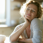cigna-therapist-newark-nj - Caring Clarity Counseling Woman Sits at a Sunlit Kitchen Table, Resting Her Chin on Her Hand and Smiling by a White Mug of Coffee.",