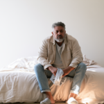 Man with Gray Beard Sits on the Edge of a Bed in a Bright, Minimalist Bedroom, Holding a Striped Cloth and Looking Thoughtful.