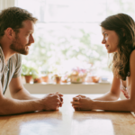 does-couples-therapy-work - Caring Clarity Counseling Man and Woman Sit Across from Each Other at a Wooden Table, Hands Clasped, in a Sunlit Kitchen with Potted Plants.
