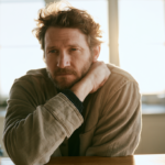 Bearded Man Sits at a Wooden Table in a Sunlit Kitchen, Resting His Chin on His Hands.