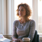 Woman with Wavy Hair Sits at a Desk by a Sunlit Window, Looking Thoughtful, with a Laptop and Coffee Cup Nearby.