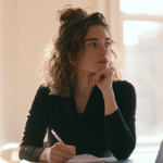 horizon-bcbs-therapist-jersey-city-nj - Caring Clarity Counseling Woman Seated at a Desk, Writing in a Notebook While Resting Her Chin, Laptop Beside Her in a Sunlit Room.