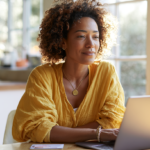 Woman with Curly Hair in a Yellow Blouse Sits at a Wooden Table with a Laptop in a Bright, Sunlit Kitchen.
