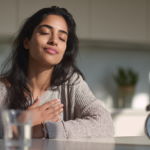 Calm Woman with Eyes Closed, Hand on Her Chest, Basking in Sunlight at a Kitchen Table with a Glass of Water and an Alarm Clock Nearby.