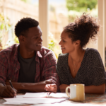 how-long-is-premarital-counseling - Caring Clarity Counseling Smiling Couple Sits at a Sunlit Kitchen Table, Writing in a Notebook with Mugs Nearby.