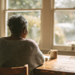 Older Person with Short Curly Gray Hair Sits at a Wooden Kitchen Table by a Sunlit Window, Facing Away.