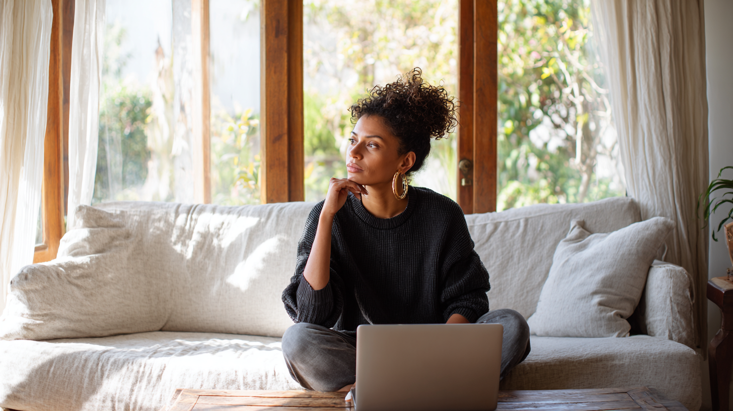 Woman with Curly Hair Sits Cross-legged on a Beige Sofa with a Laptop, Gazing Thoughtfully by Sunny Windows.