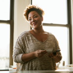 Smiling Woman in a Sunlit Kitchen Holds a Credit Card and Smartphone by the Counter Near a Large Window in a Casual Striped Sweater of Gray Tones.