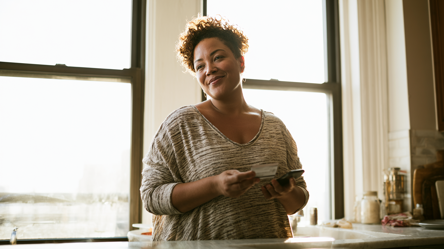 Smiling Woman in a Sunlit Kitchen Holds a Credit Card and Smartphone by the Counter Near a Large Window in a Casual Striped Sweater of Gray Tones.