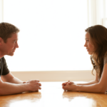 Man and Woman Sit Across from Each Other at a Wooden Table, Hands Clasped, Engaged in Conversation in a Bright Room.