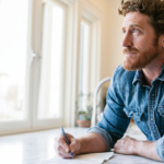 Man in a Blue Denim Shirt Writes in a Notebook at a Bright, Sunlit Kitchen Counter by Large Windows.