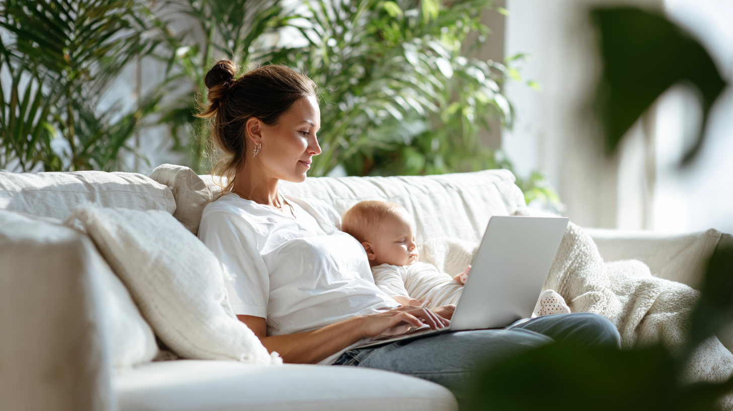 Mother Relaxing on a Light Sofa with Her Baby on Her Lap, Typing on a Laptop in a Bright Living Room.