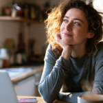 Woman with Curly Hair Sits at a Kitchen Table, Resting Her Chin on Her Hand, Smiling Softly at a Laptop Nearby.