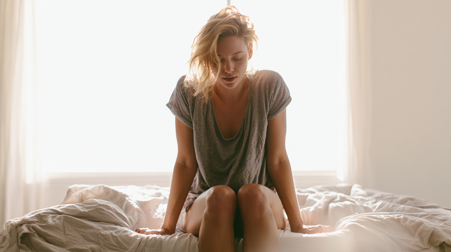Woman Sitting on a Bed in a Sunlit Room, Head Bowed, Wearing a Loose Gray T‑shirt, Knees Drawn Up on the Duvet.