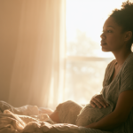 pregnancy-depression - Caring Clarity Counseling Pregnant Woman Sitting on a Sunlit Bed, Cradling Her Belly and Gazing off Thoughtfully.
