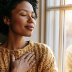 psychological-trauma - Caring Clarity Counseling Woman with Short Curly Hair, Wearing a Chunky Tan Sweater, Eyes Closed, Hand on Her Chest by a Sunlit Window.