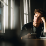 Young Man Sits at a Desk by a Window, Resting His Head on One Hand While Looking at His Phone in Front of a Laptop.