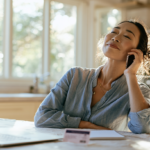 Smiling Woman Sits at a Sunlit Kitchen Table, Talking on Her Smartphone with a Laptop Open Nearby and a Notebook in Front of Her.