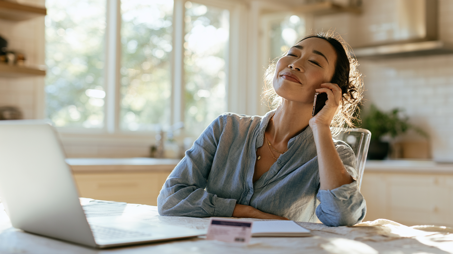Smiling Woman Sits at a Sunlit Kitchen Table, Talking on Her Smartphone with a Laptop Open Nearby and a Notebook in Front of Her.