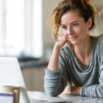 Woman in a Gray Long-sleeve Top Rests Her Chin on Her Hand While Looking at a Laptop on a Kitchen Table.