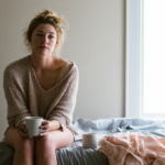 Young Woman Sits on a Bed in a Soft Knit Sweater, Holding a Mug As Natural Light Pours in from a Window to the Right.