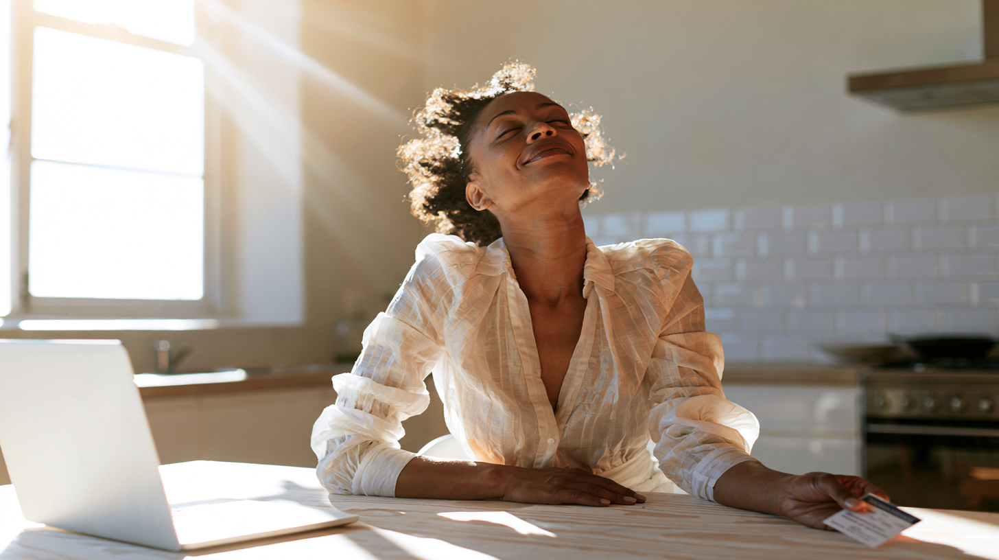 Woman in a White Blouse Sits at a Sunlit Kitchen Table with a Laptop, Smiling at the Bright Window.