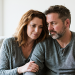 Couple Sitting Closely on a Light Sofa, Holding Hands and Sharing a Quiet, Supportive Moment at Home.