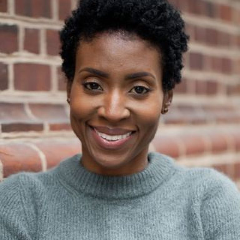 Smiling Black woman with short curly hair wearing a gray knit sweater, leaning against a brick wall in the background.