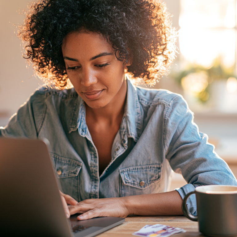Woman with curly hair in a denim shirt works on a laptop at a sunlit kitchen table, a mug nearby.