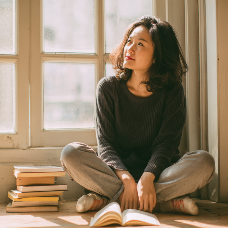 Woman sits cross‑legged on a wooden floor by a sunlit window, reading an open book with a stack of books beside her