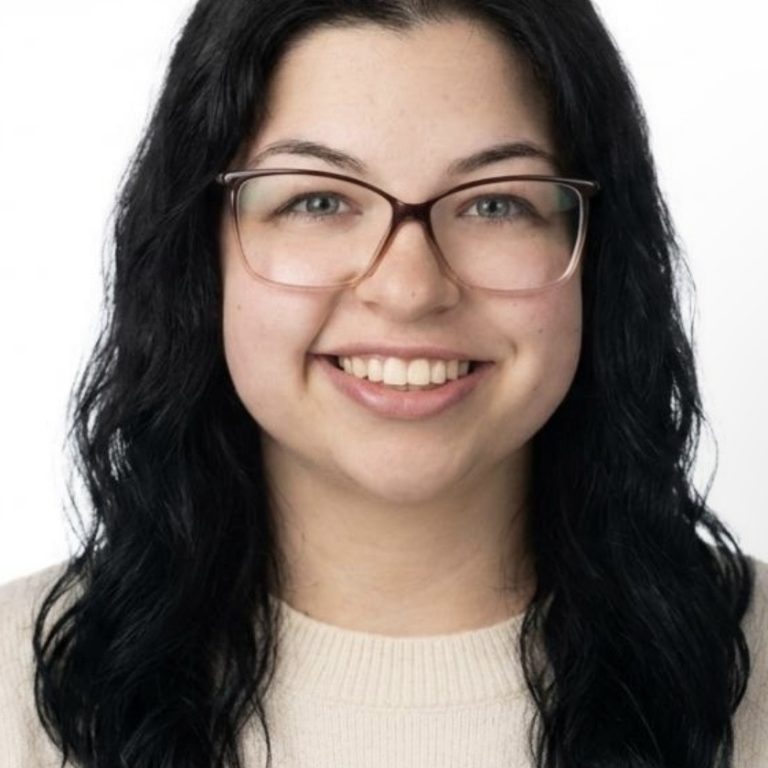 Smiling woman with long dark hair and pink glasses, wearing a beige sweaters in a head-and-shoulders portrait.