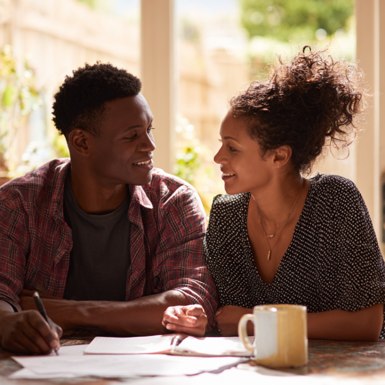 Smiling couple sits at a sunlit kitchen table, writing in a notebook with mugs nearby.