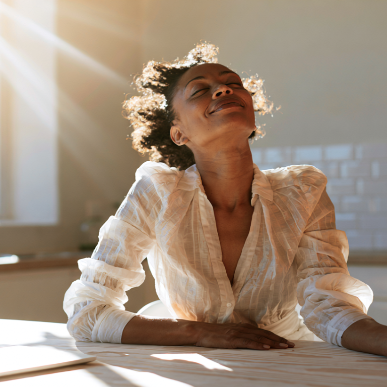 Woman in a white blouse sits at a sunlit kitchen table with a laptop, smiling at the bright window.