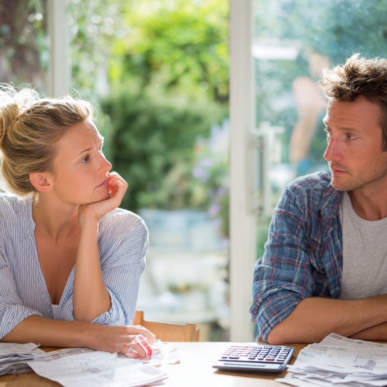 Couple sits at a sunlit kitchen table, surrounded by bills and a calculator, looking at each other pensively.