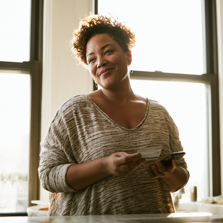 Smiling woman in a sunlit kitchen holds a credit card and smartphone by the counter near a large window in a casual striped sweater of gray tones.