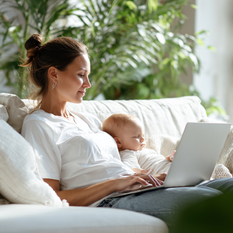 Mother relaxing on a light sofa with her baby on her lap, typing on a laptop in a bright living room.