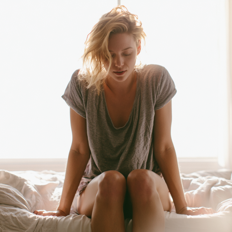Woman sitting on a bed in a sunlit room, head bowed, wearing a loose gray t‑shirt, knees drawn up on the duvet.