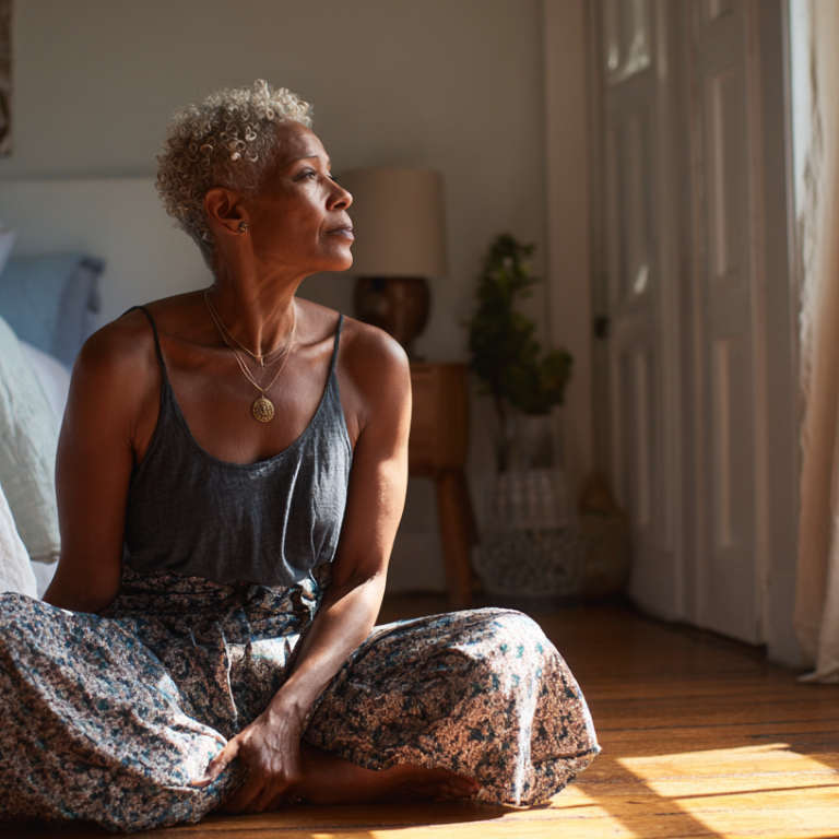Older woman with short gray hair sits cross-legged on the floor in a sunlit bedroom, gazing to the right, wearing a gray tank top and floral skirt.