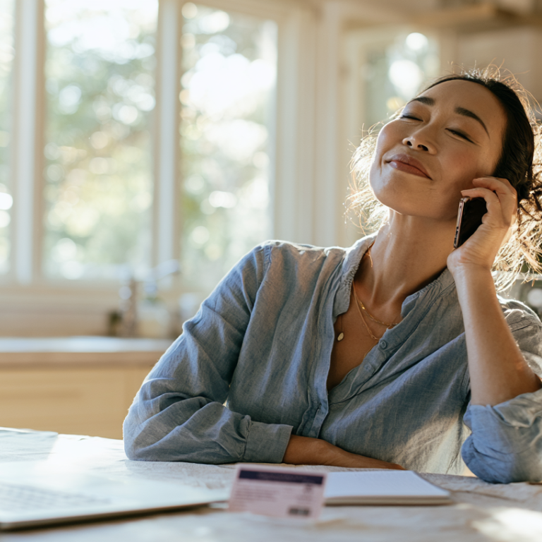 Smiling woman sits at a sunlit kitchen table, talking on her smartphone with a laptop open nearby and a notebook in front of her.