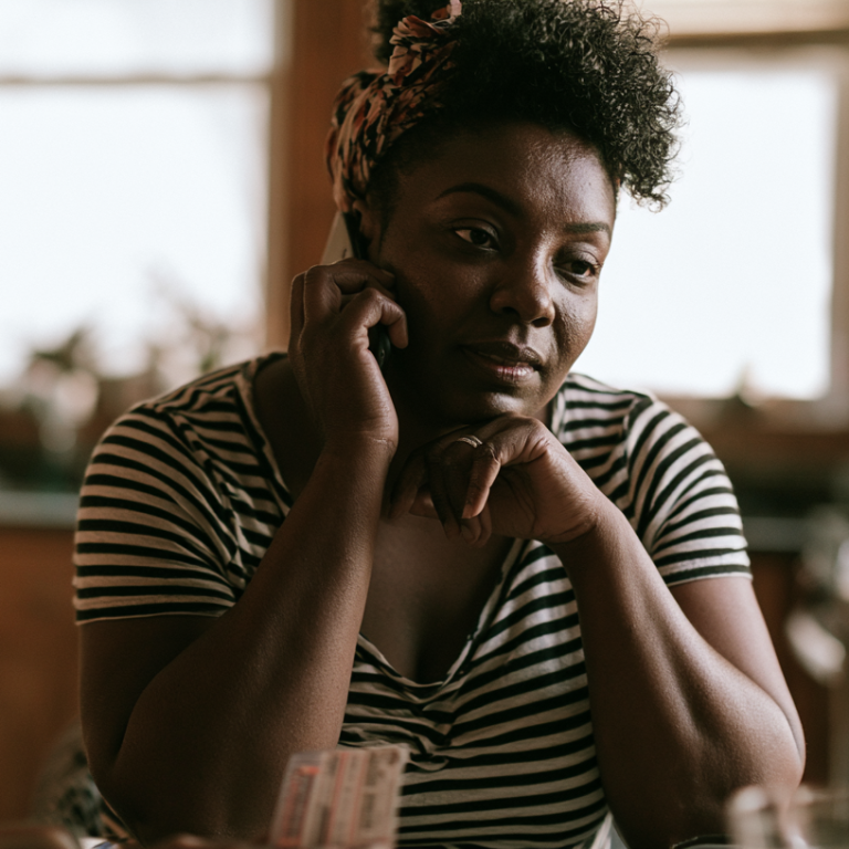 Woman in a black-and-white striped shirt talks on a cell phone, resting her chin on her hands with a thoughtful expression indoors at home.